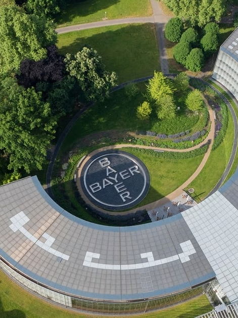 Aerial view of a U-shaped building with a circular garden in the center, featuring the Bayer logo. Surrounded by trees and a pathway.