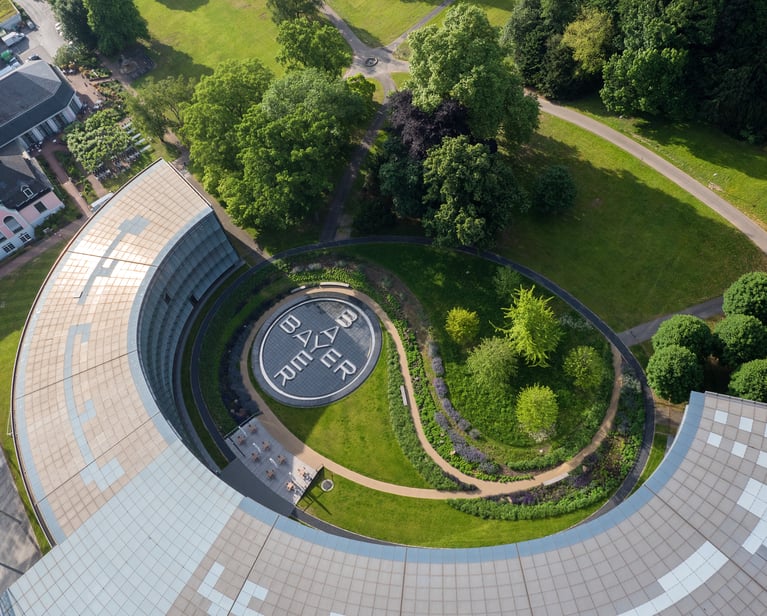 Aerial bird's-eye view of the curved Bayer headquarters building. In the center of the green, landscaped courtyard, a large circular pavement feature prominently displays the iconic Bayer cross logo.