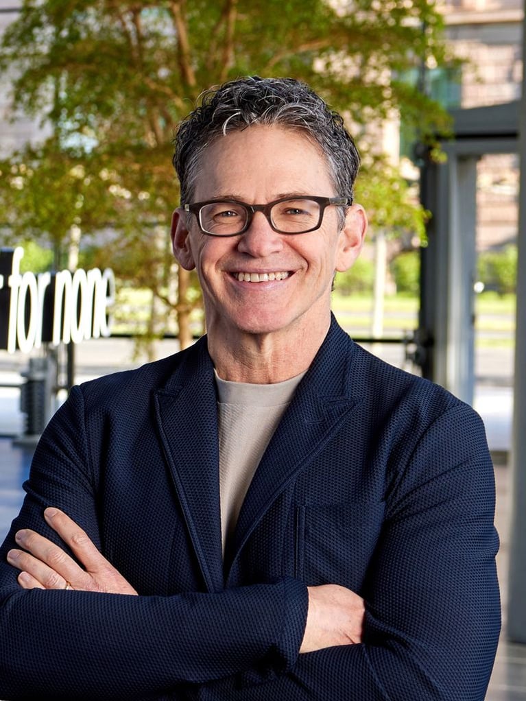 Portrait of Bayer CEO Bill Anderson smiling with arms crossed, wearing glasses and a dark blue jacket, with a blurred outdoor background.