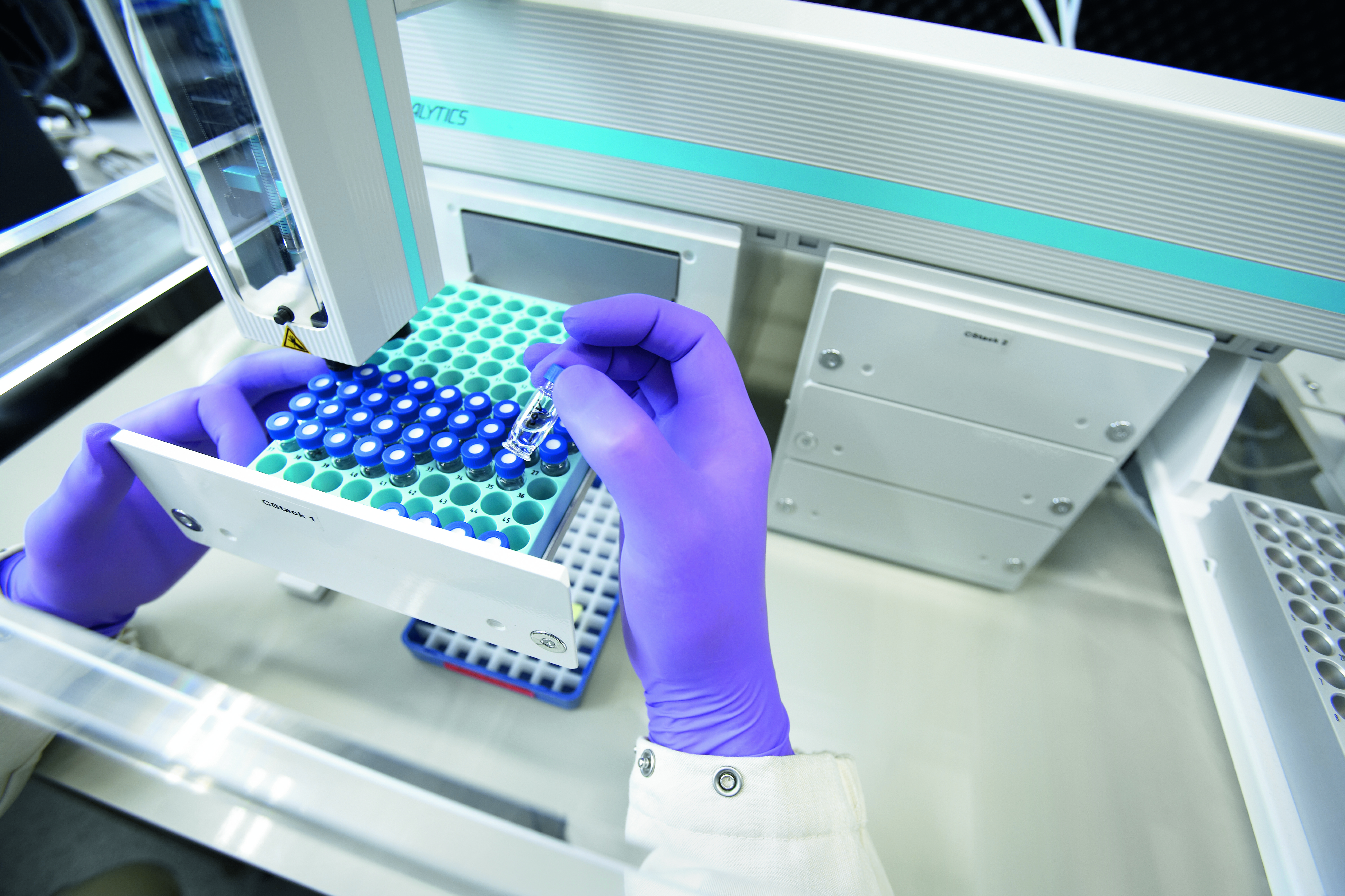 A lab technician with gloved hands handling sample vials near automated laboratory equipment.