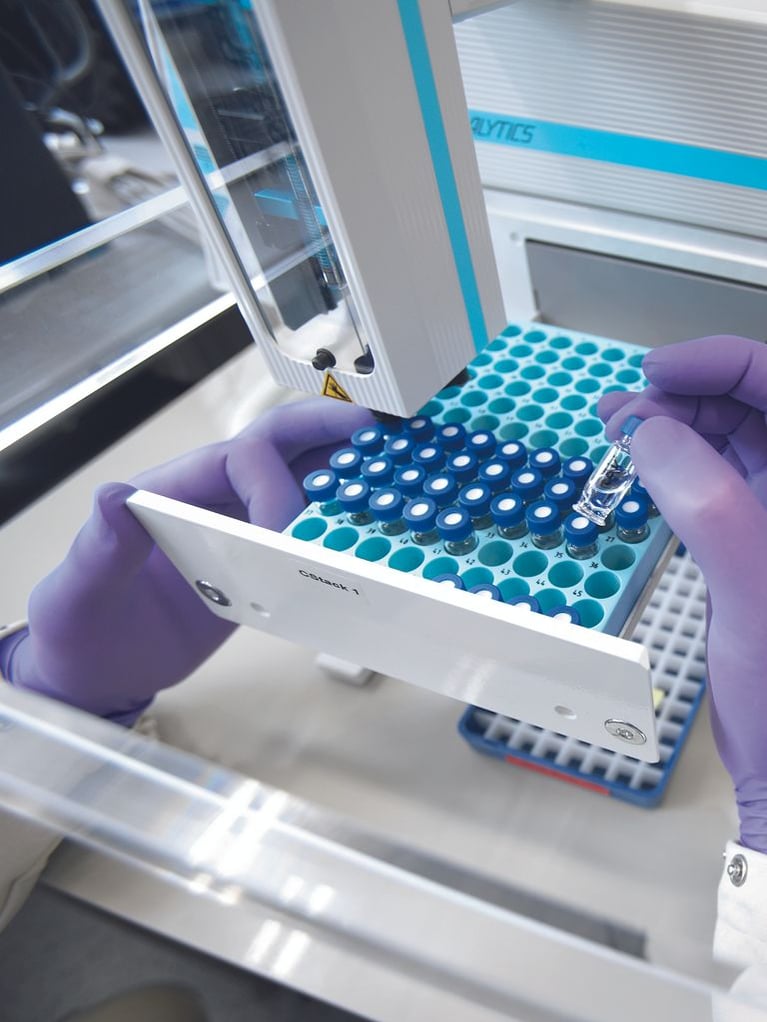 A lab technician with gloved hands handling sample vials near automated laboratory equipment.