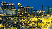 Imagem de uma grande instalação industrial com tubulações, torres metálicas e estruturas iluminadas à noite, representando a fase de reconstrução e crescimento da Bayer durante o período do “milagre econômico” entre 1951 e 1974.