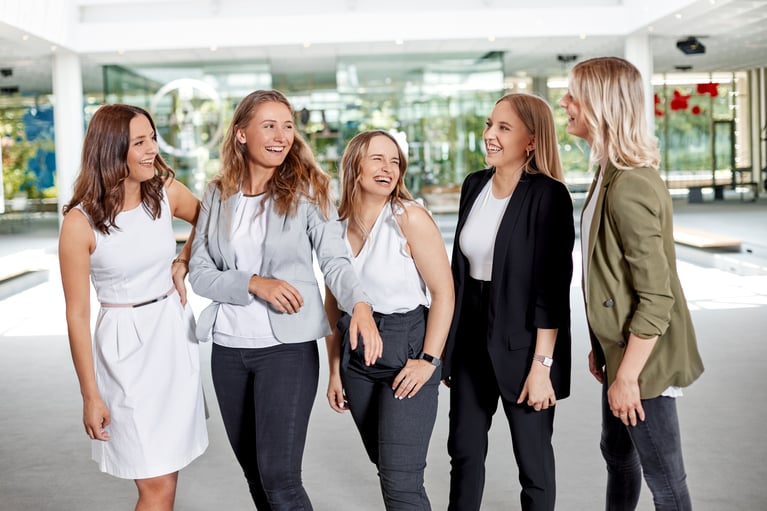 A group of five women standing together indoors, smiling and laughing, dressed in professional attire.