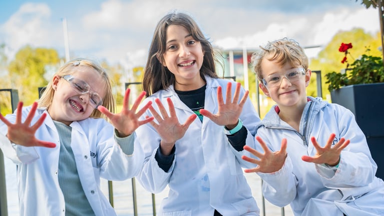 Three laughing children in white lab coats holding up their red-stained palms to the camera.