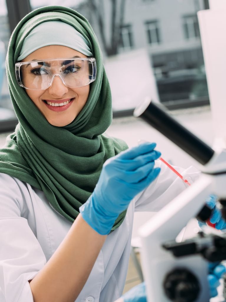 A smiling female scientist wearing a green hijab, safety goggles, and a white lab coat looks to the side while working with a microscope and pipette in a laboratory.