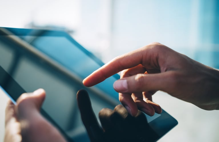 Close-up of a person's hands holding a digital tablet outdoors, with a finger touching the glossy screen reflecting light and shadows.