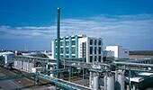 Imagem de uma instalação industrial moderna com edifícios brancos e estruturas metálicas, incluindo uma torre alta e tubulações, sob um céu azul, representando a fase de transformação e globalização da Bayer entre 1988 e 2001.