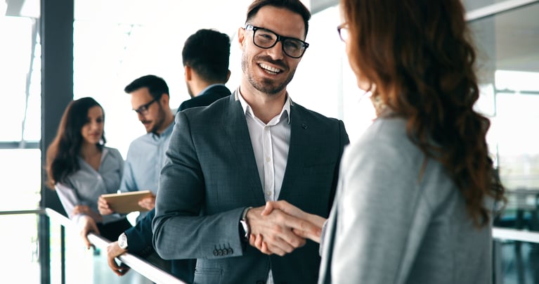 Two professionals shaking hands in a modern office setting, with two colleagues conversing in the background.