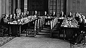 Imagem histórica em preto e branco mostrando uma grande sala com várias pessoas sentadas em longas mesas durante uma reunião formal, representando o período de dissolução da I.G. Farben e o reestabelecimento da Bayer entre 1945 e 1951.