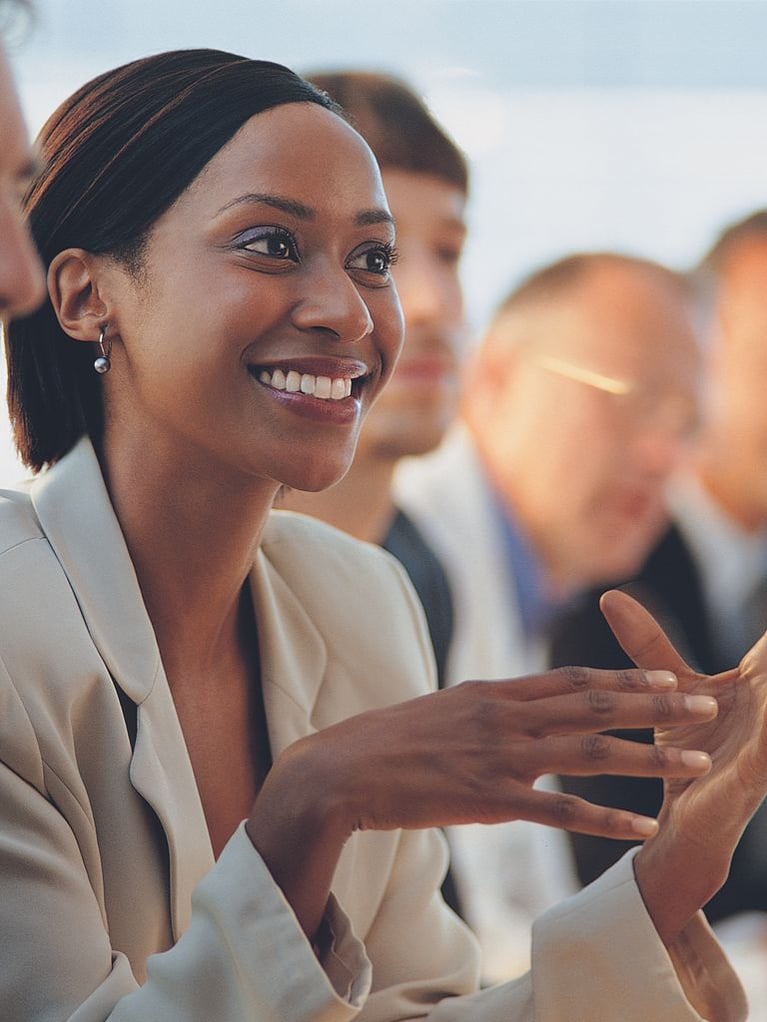 A smiling businesswoman wearing a light beige blazer gestures with her hands while speaking in a meeting, with colleagues blurred in the background.