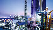Imagem de uma instalação industrial moderna com estruturas metálicas, tubulações e plataformas elevadas, iluminadas com luz artificial, incluindo uma pessoa com capacete de segurança operando equipamentos, representando a reorganização da Bayer entre 2001 e 2010.