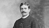 Retrato histórico em preto e branco de uma pessoa do início do século XX, vestindo uniforme formal escuro com gola alta, representando a fase de expansão internacional da Bayer entre 1881 e 1914.