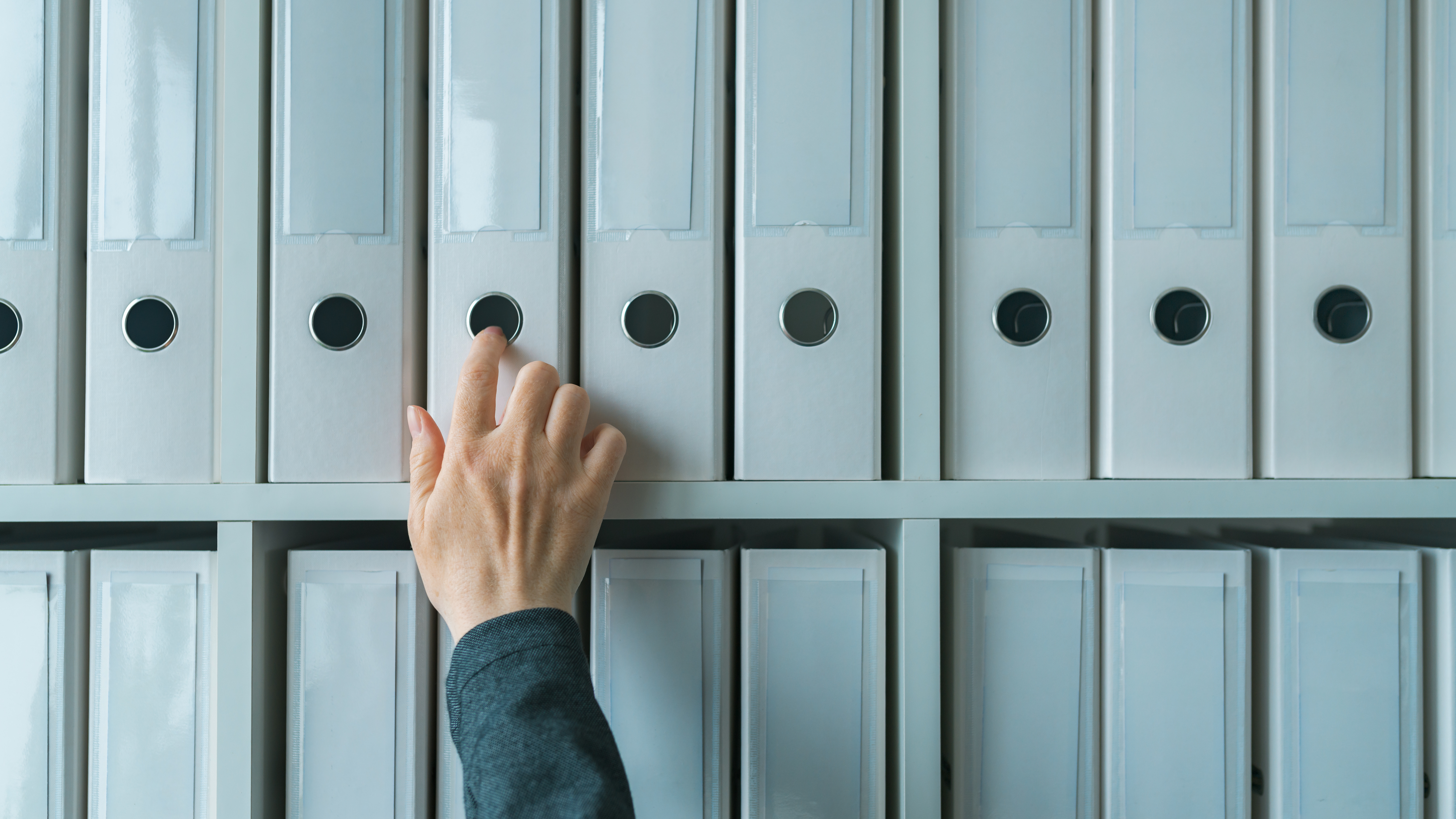 A hand reaching for a white ring binder on a shelf filled with similar binders.