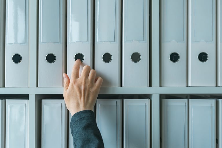 A hand reaching for a white ring binder on a shelf filled with similar binders.