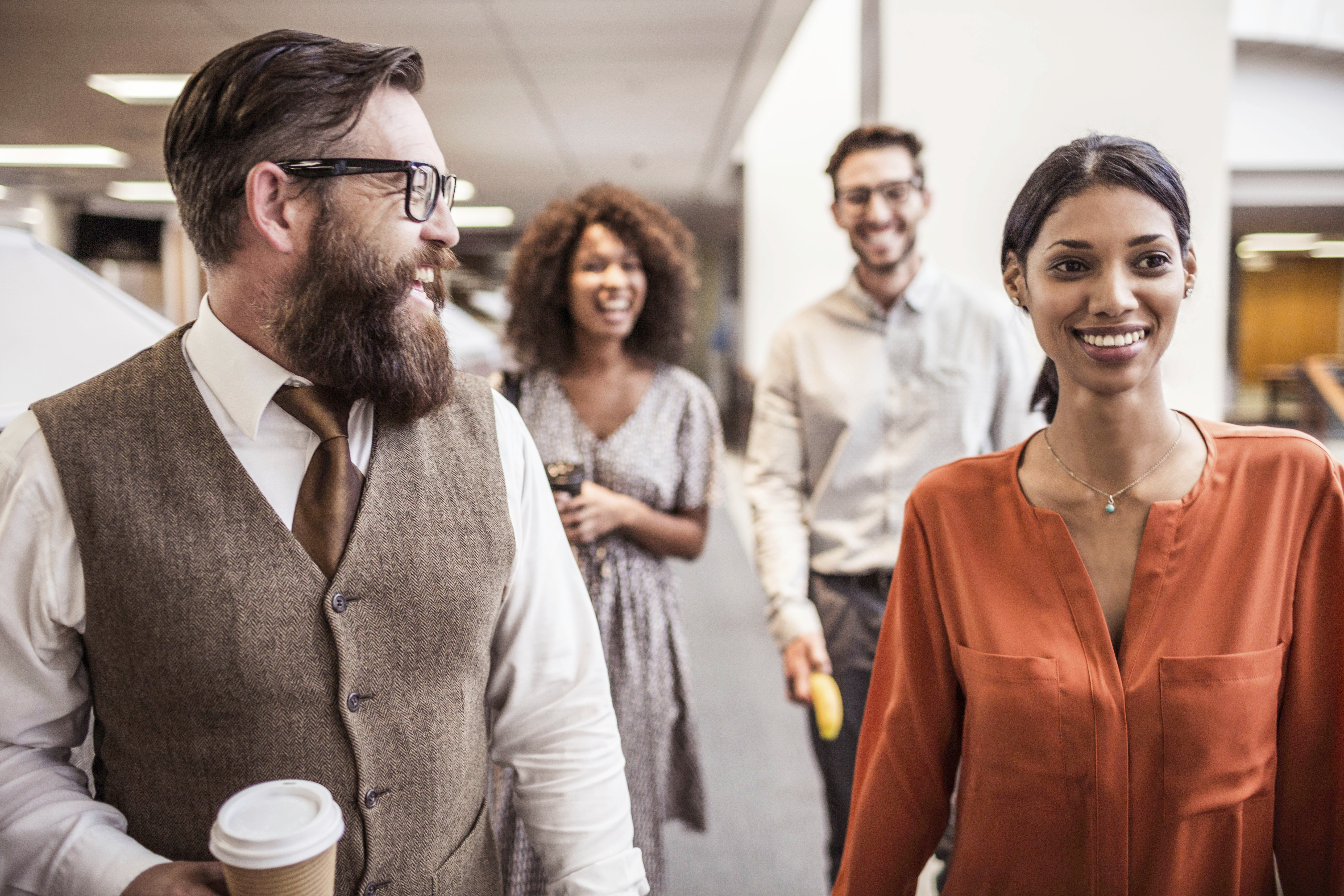 A bearded man in the foreground is smiling and holding a coffee cup, while other laughing people walk through a hallway in the background.