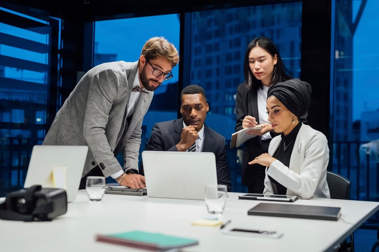 Four professionals gather around a laptop at a conference table in an office at night. A man leans over the screen, another sits looking thoughtfully, a woman takes notes, and a woman wearing a black turban gestures while looking at the display.