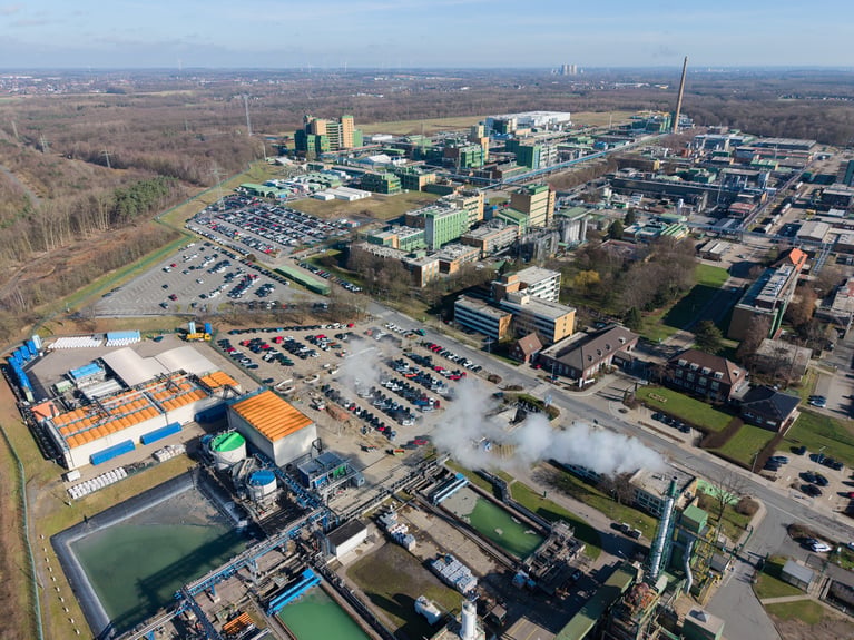 Aerial view of a large industrial plant with parking lots, processing tanks, and steam rising beside wooded land.